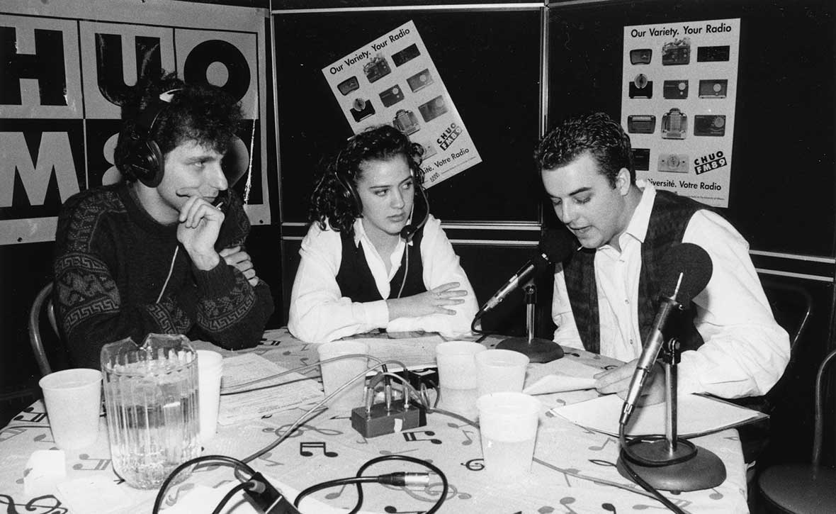 Black-and white photo of three CHUO-FM radio hosts, sitting at a table on which we see microphones, glasses and a pitcher of water. From left to right: Vincent Veilleux, Claudia Fillion and Stefan Psenak