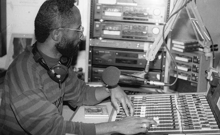 A radio host sits at a radio studio mixing table with headphones around his neck.