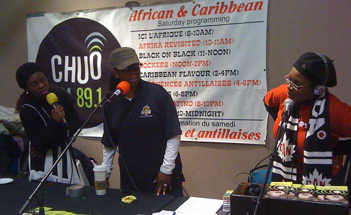 Denise Moore, Adrienne Coddett and Sarah Onyango , three radio hosts for an African and West Indian program, stand in front of a table, each with a microphone. In the background, a CHUO poster lists the Saturday program line-up. 