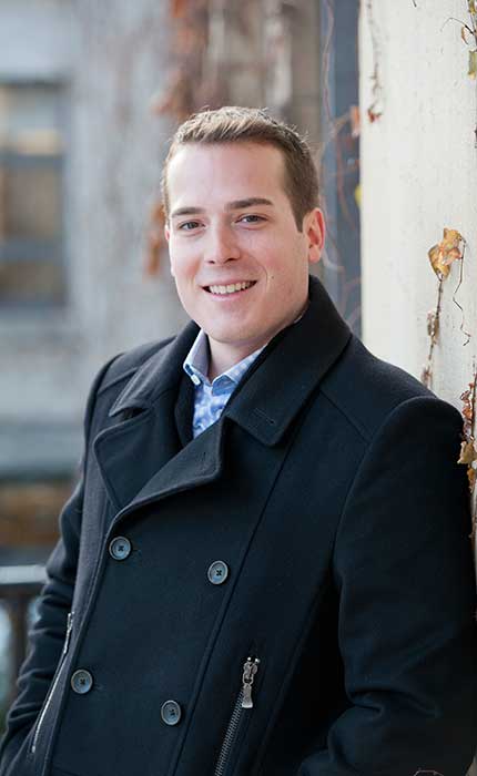 Mathieu Fleury, smiling and standing outside of Tabaret Hall as he leans against a pillar.  He is wearing a black buttoned coat. Photo taken during the day.