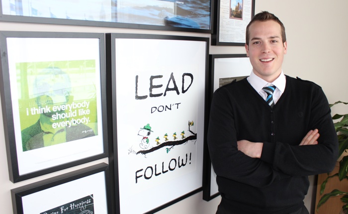 Mathieu Fleury, standing, wears a black v-neck sweater on a white shirt and a blue, black and grey-striped tie. He is standing in front of some posters, one of which reads, “Lead, don’t follow”.