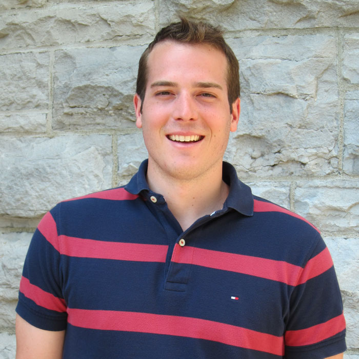 Front view of Mathieu Fleury, smiling. He is wearing a navy blue polo shirt with red stripes. A light-grey stone wall is in the background. Photo taken during the day.
