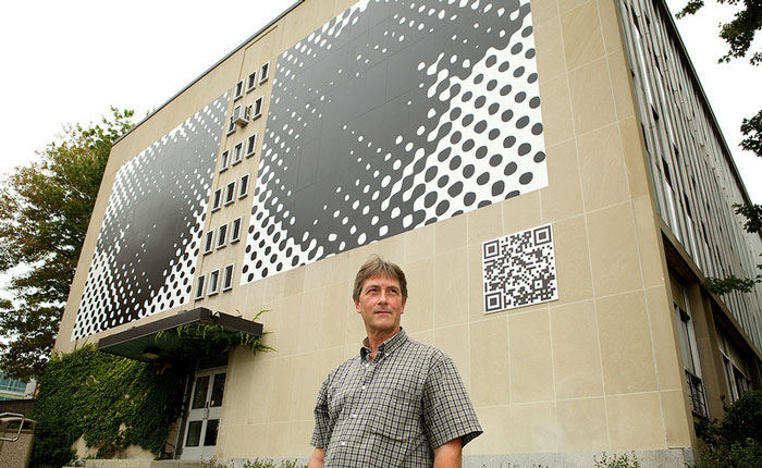 Artist Steven Boyd looking to the right (his left). He is standing in front of the work, which is photographed from a ground angle. Photo taken during the day, in summer